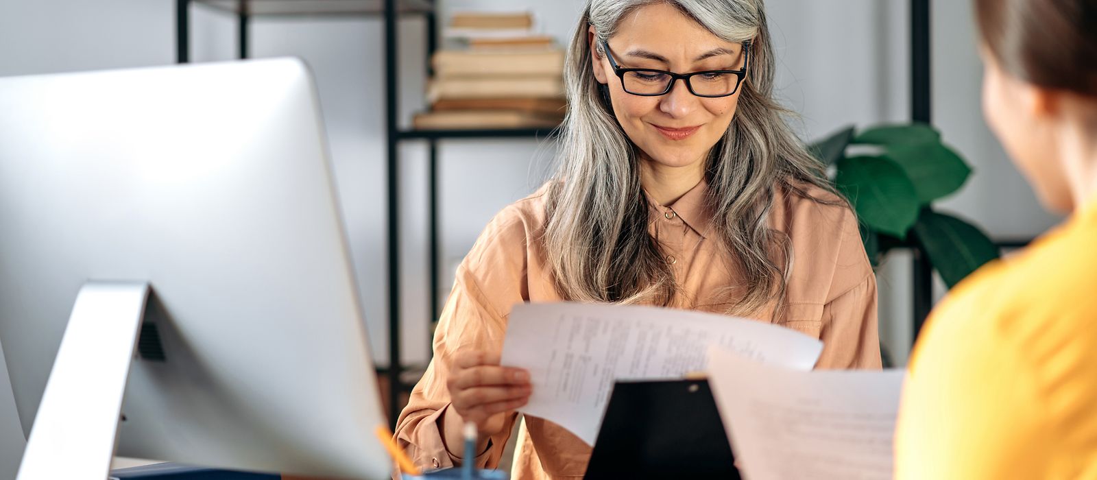 Frau mit Brille sitzt vor ihrem Computer und guckt auf ein Blatt Papier