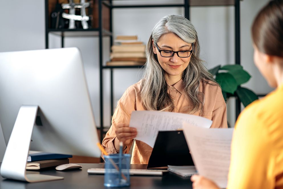 Frau mit Brille sitzt vor ihrem Computer und guckt auf ein Blatt Papier